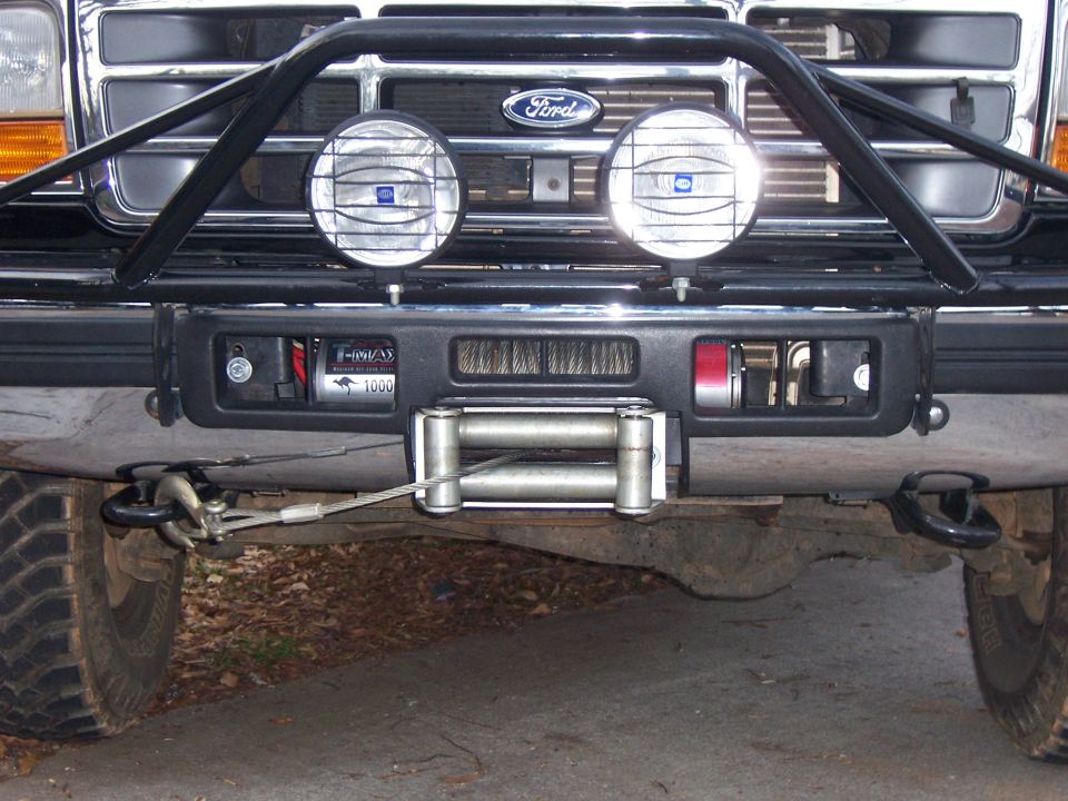 Front bumper of a dark-colored Ford Bronco with a winch, two large auxiliary lights, and a brush guard