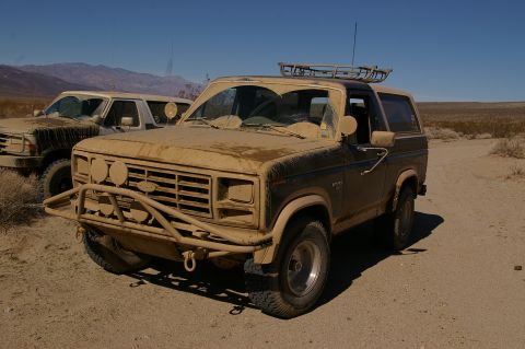 1986 Ford Bronco - Two dusty off-road vehicles sit idle on a dirt road under a clear blue sky, with mountains in the distance.