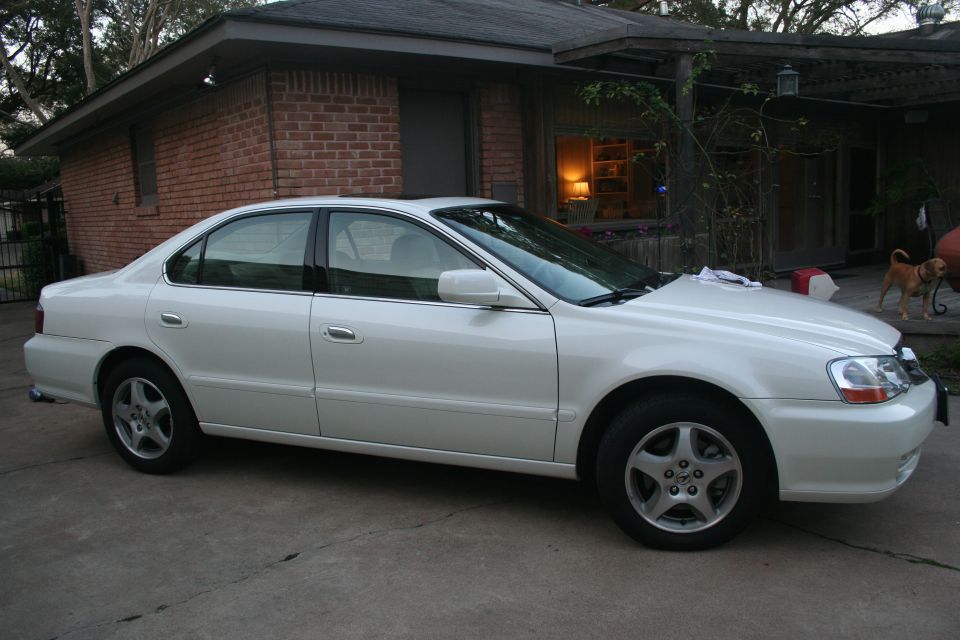 White Acura TL sedan parked in a driveway in front of a brick house, side view
