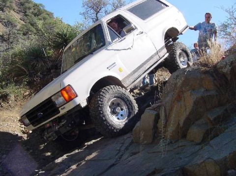 1987 Ford Bronco - A white 1987 Ford Bronco navigates a challenging rocky terrain on a sunny day.