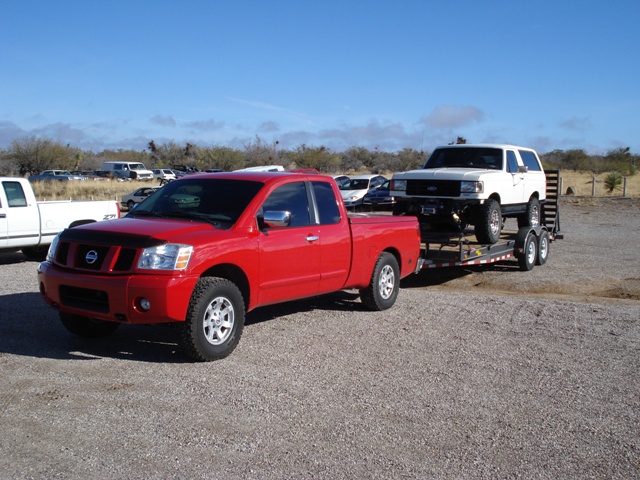 Red Nissan Titan pickup towing a trailer loaded with a lifted white Ford Bronco on gravel