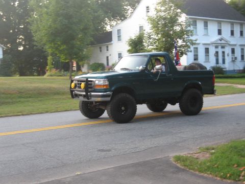 1996 Ford Bronco - A dark green Ford Bronco is seen driving on a road with houses in the background.