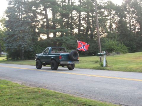 1996 Ford Bronco - Three-quarter rear view of a lifted dark green Ford Bronco with its hardtop removed, driving down a two-lane rural road lined with pine tre…