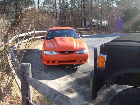 1995 Ford Mustang - An orange Ford Mustang is parked on a dirt road next to a black truck.