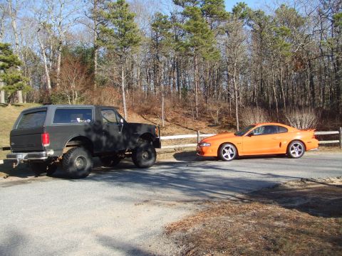 1995 Ford Mustang - An orange 1995 Ford Mustang coupe is parked next to a lifted black SUV.
