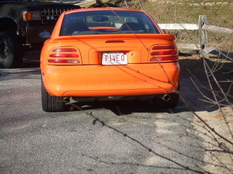 1995 Ford Mustang - The rear of an orange 1995 Ford Mustang GT is visible, parked next to a dark-colored SUV.