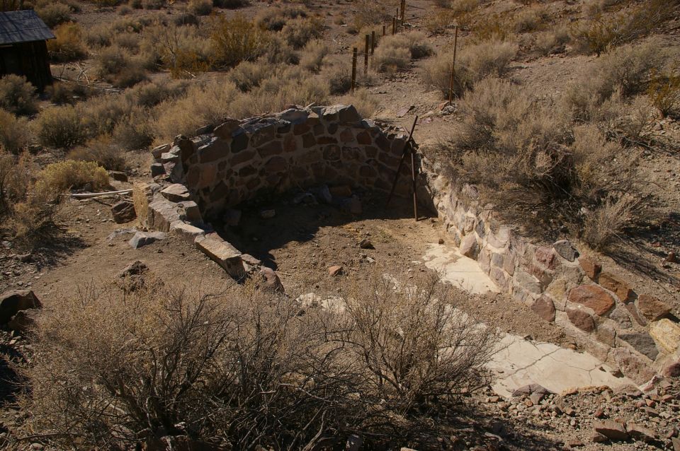 Stone foundation ruins in a dry, brushy landscape with a small wooden structure in the background