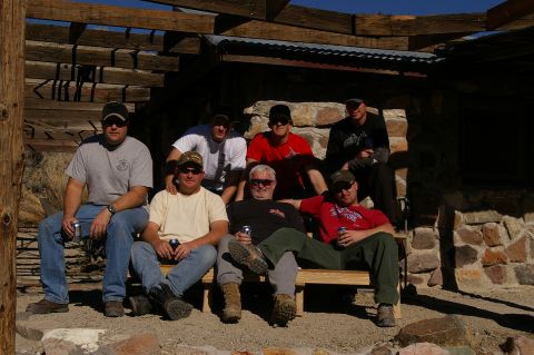 1986 Ford Bronco - Seven men are gathered outdoors, seated on a wooden bench in front of a stone structure with a wooden overhang.