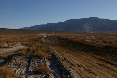 1986 Ford Bronco - A 1986 Ford Bronco kicks up dust as it travels along a dirt road in a vast desert landscape under a clear blue sky.