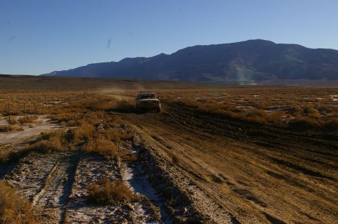 1986 Ford Bronco - A vehicle navigates a dusty trail through a barren, arid landscape under a clear blue sky.