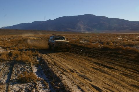 1986 Ford Bronco - A 1980s Ford Bronco navigates a muddy track through a dry, arid terrain with mountains in the background.