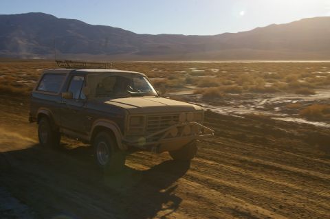 1986 Ford Bronco - A 1986 Ford Bronco kicks up dust as it drives through a desert landscape at sunset.