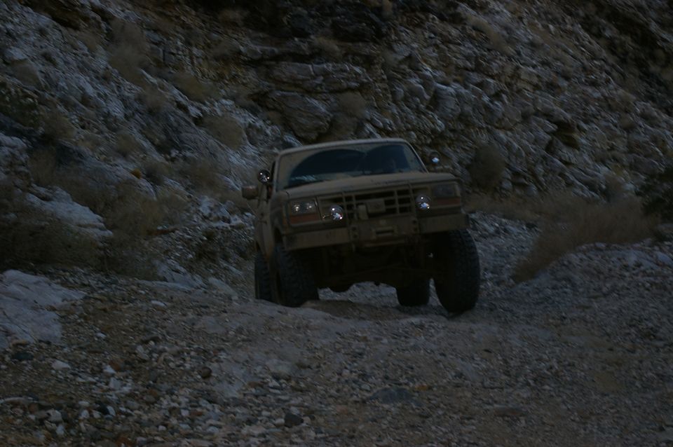 Tan Ford Bronco climbing a steep, rocky incline. Headlights are on