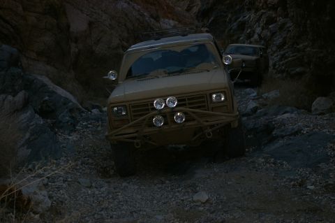 1986 Ford Bronco - Two off-road SUVs navigate a narrow, rocky canyon trail in the desert.