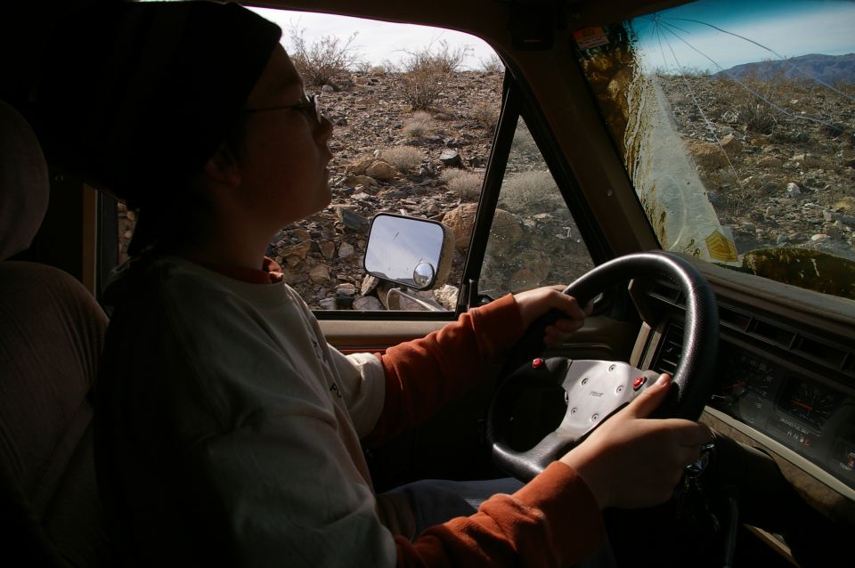 1986 Ford Bronco interior, driver's hands on steering wheel, cracked windshield showing desert landscape