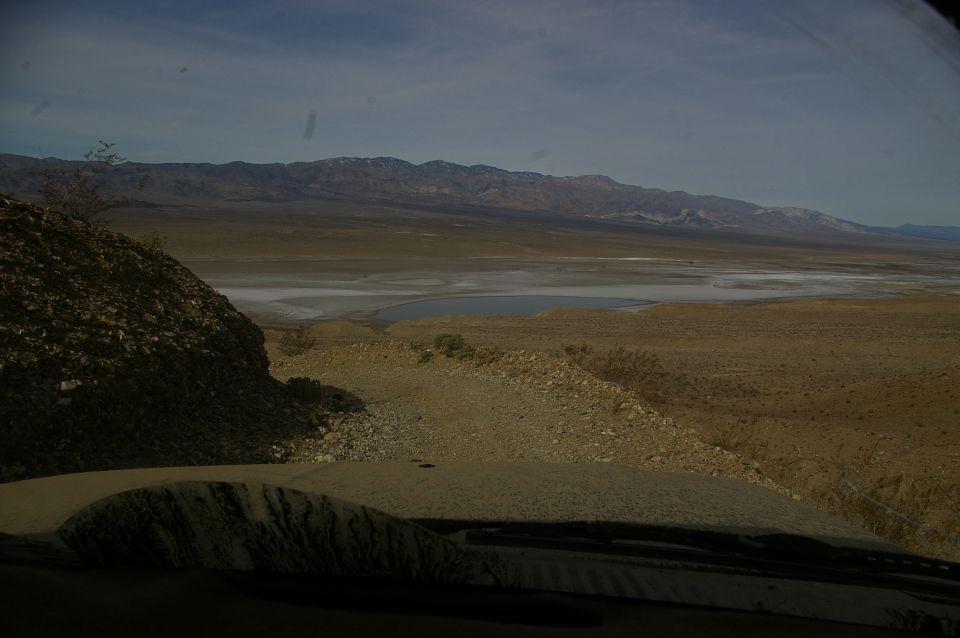 View from inside a vehicle looking out at a vast desert landscape with mountains in the distance and a dry lake bed