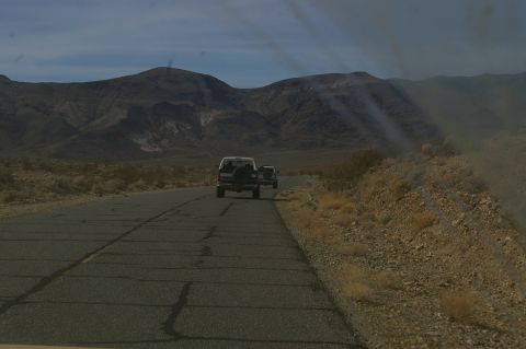1986 Ford Bronco - Two SUVs travel down a desolate desert highway under a partly cloudy sky.