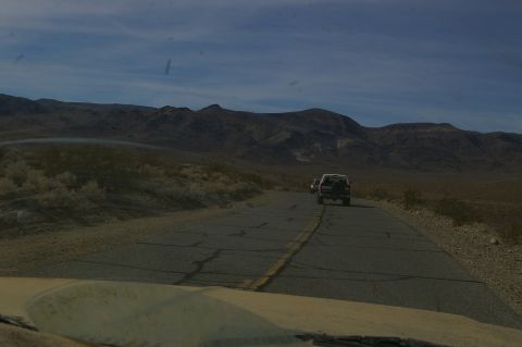1986 Ford Bronco - Two SUVs travel down a cracked asphalt road through a barren, mountainous desert landscape under a blue sky.