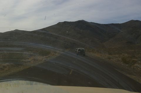 1986 Ford Bronco - Two SUVs travel on a dusty road through a barren, hilly desert environment under a cloudy sky.