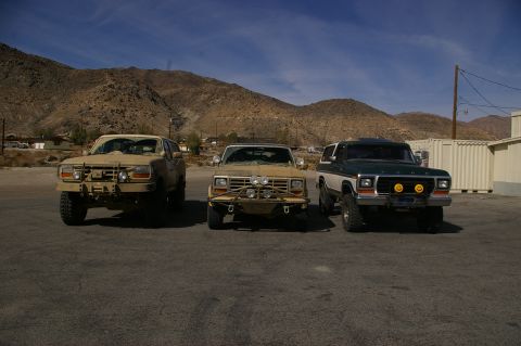 1986 Ford Bronco - Three SUVs, two tan and one two-tone green and white, are parked in a lot with mountains behind them.