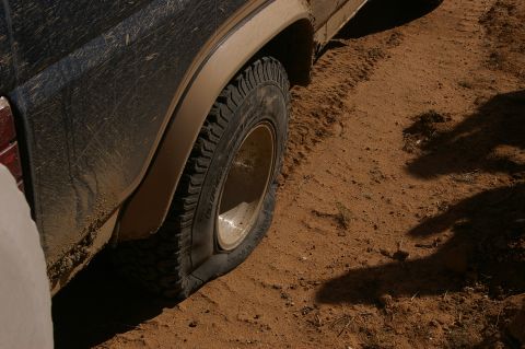 1986 Ford Bronco - A vehicle's tire is flat in the sand, with mud splattered on the body.