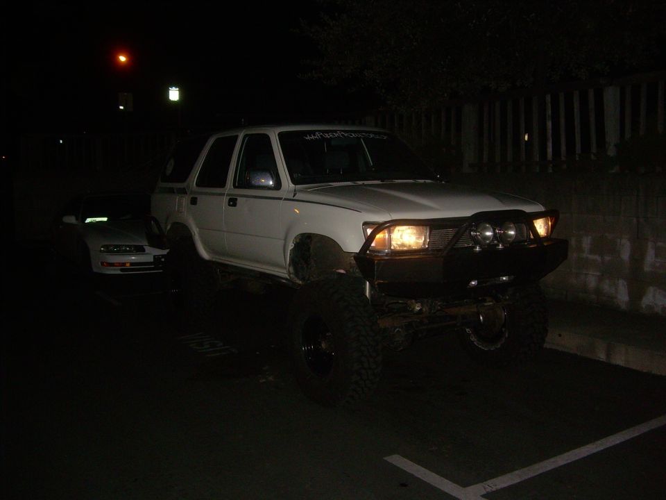 White SUV with large tires and a bull bar, parked at night. Another car is visible behind it