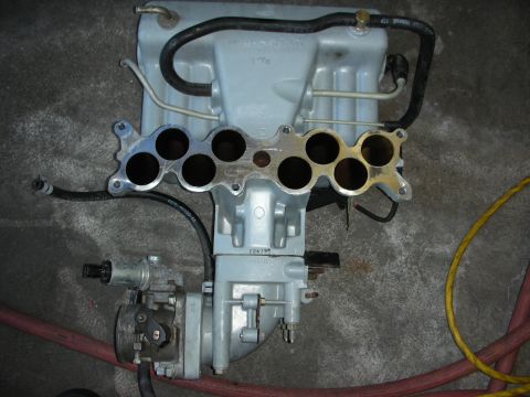 1998 Ford Crown Victoria - grey aluminum intake manifold with eight open runner ports and an attached throttle body sitting on a concrete floor.