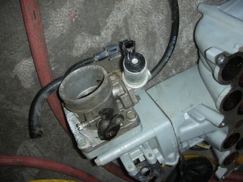 1998 Ford Crown Victoria - throttle body with an idle air control valve and electrical connector mounted to a grey intake manifold on the floor.