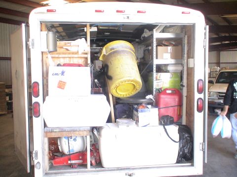 1993 Ford C-350 (Centurion Conversions) - The rear of a white cargo trailer is open, revealing a cluttered interior with storage shelves, containers, and a large yellow barrel.