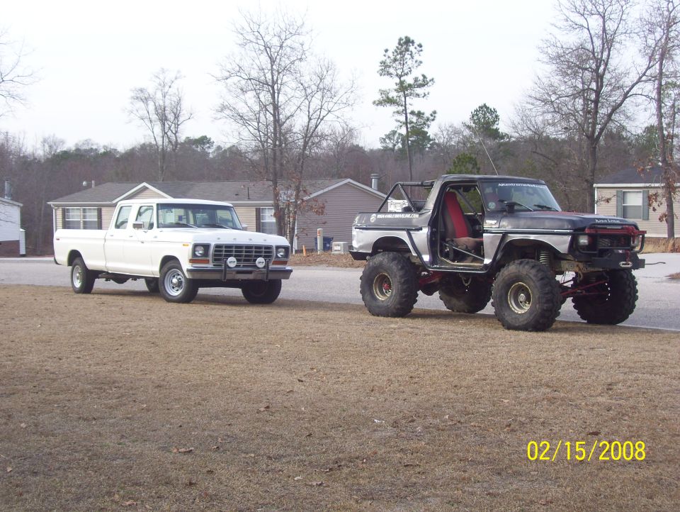 Two Ford trucks, one white crew cab and one lifted silver Bronco, parked on a dirt road