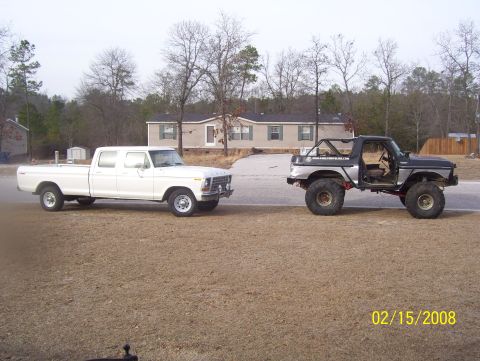 1978 Ford Bronco - A lifted Bronco-like vehicle and a white crew cab pickup truck are parked on a dirt lot with a house in the background.