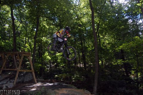 1976 Toyota Land Cruiser - A mountain biker soars through the air during a downhill jump in a forest setting.