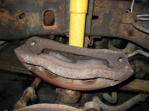 1995 Ford C-350 (Centurion Conversions) - Close-up of a rusty front brake caliper and rotor assembly, with a yellow shock absorber in the background.
