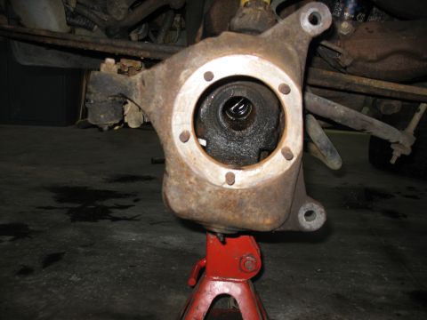 1995 Ford C-350 (Centurion Conversions) - Close-up of a rusty front axle spindle, supported by a red jack stand, with the vehicle's undercarriage visible above.