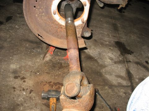 1995 Ford C-350 (Centurion Conversions) - Close-up of a rusty driveshaft U-joint and brake rotor assembly, supported by an orange jack stand.