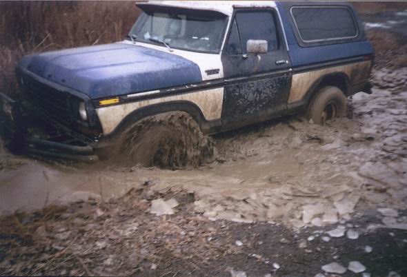 Blue and white Ford Bronco stuck in mud, front wheel deep, side view