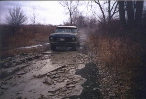 1978 Ford Bronco - A dark pickup truck navigates a challenging muddy road, its front grille and headlights visible.