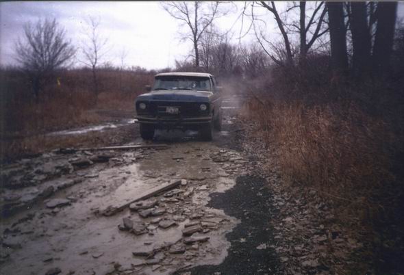 Front view of a dark 1970s pickup truck on a muddy, rutted road