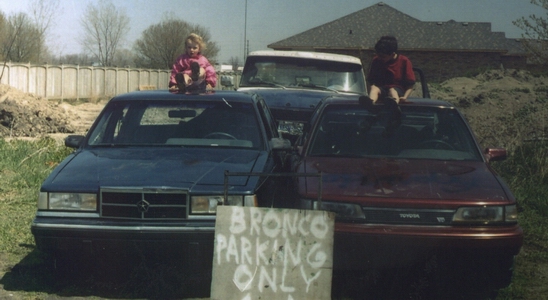 Two kids sit on hoods of a dark blue and a maroon sedan behind a hand-painted 'Bronco Parking Only' sign.