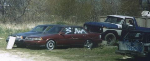 1978 Ford Bronco - Multiple vehicles are parked in an outdoor setting, with a red sedan showing white markings on its side.