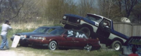 1978 Ford Bronco - A blue and white 1978 Ford Bronco is precariously perched atop a maroon sedan in a junkyard scene.