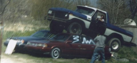 1978 Ford Bronco - A truck is precariously balanced on top of a sedan, with graffiti on the sedan's doors.