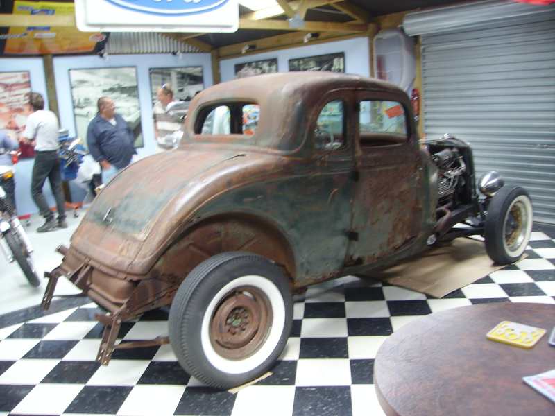 Rusty hot rod coupe with exposed engine and whitewall tires, parked indoors on a checkered floor