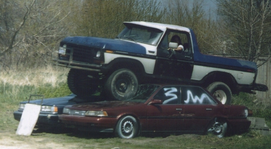 Blue and white 1978 Ford Bronco, front-mounted grille guard, on top of a maroon sedan and a dark sedan