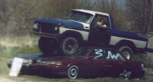Blue and white pickup truck crushing a red sedan with white graffiti on window