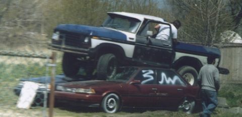 1978 Ford Bronco - A Ford F-Series truck is precariously stacked on top of a sedan in an outdoor setting.