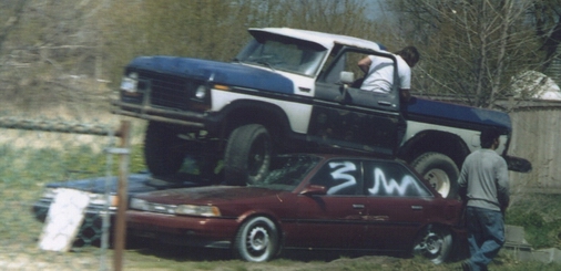 Ford F-Series truck balanced on a maroon sedan, with two people visible