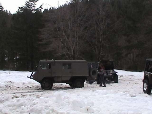 Olive drab military truck with spare tire on rear, parked in snow next to two Jeeps