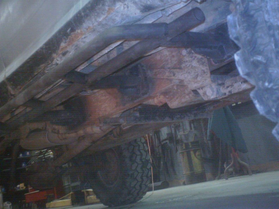 Underside of a rusty truck frame showing leaf springs, exhaust pipe and crossmembers