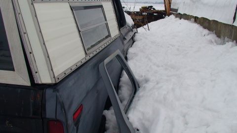 1979 Ford Bronco - A pickup truck with a camper shell sits partially buried in snow, its door open.